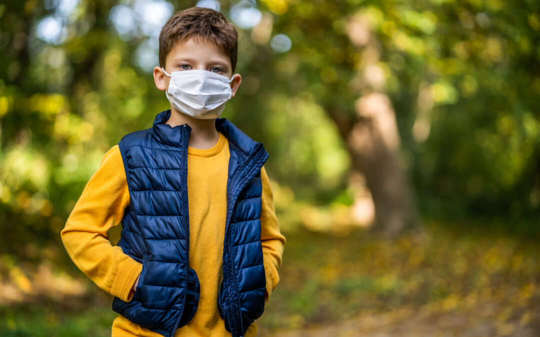 Pictured is a young boy in a face mask. He is wearing a yellow shirt, blue puffer jacket and standing in the forest.